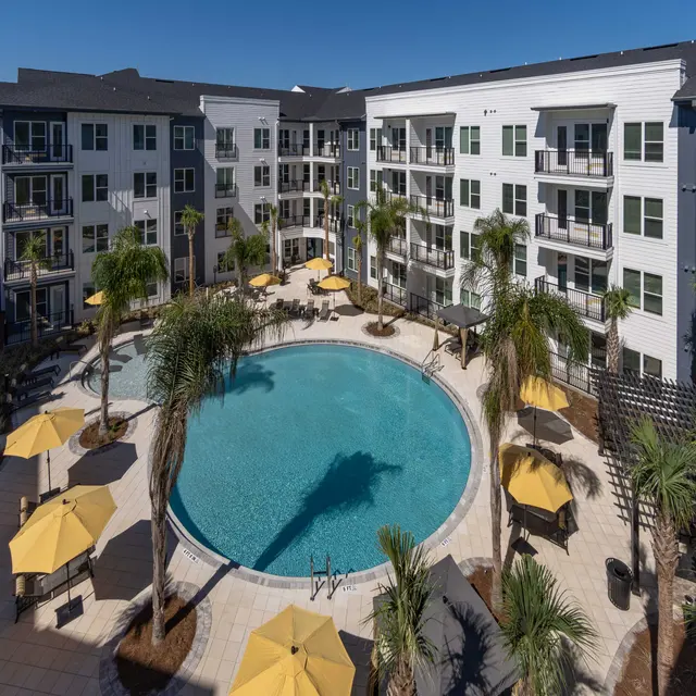 Aerial view of a modern apartment complex featuring a circular swimming pool surrounded by palm trees and sun umbrellas.