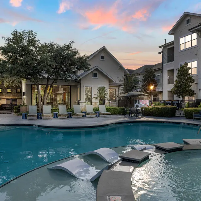 A serene apartment pool area with lounge chairs around a clear blue pool, surrounded by trees and modern buildings under a colorful sunset.