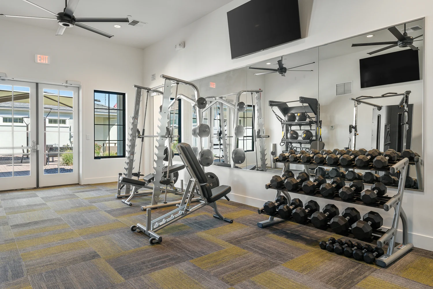 A modern gym interior featuring weightlifting equipment including a bench press, dumbbells rack, and mirrors. The space is bright with natural light coming from large windows and has a stylish carpet.