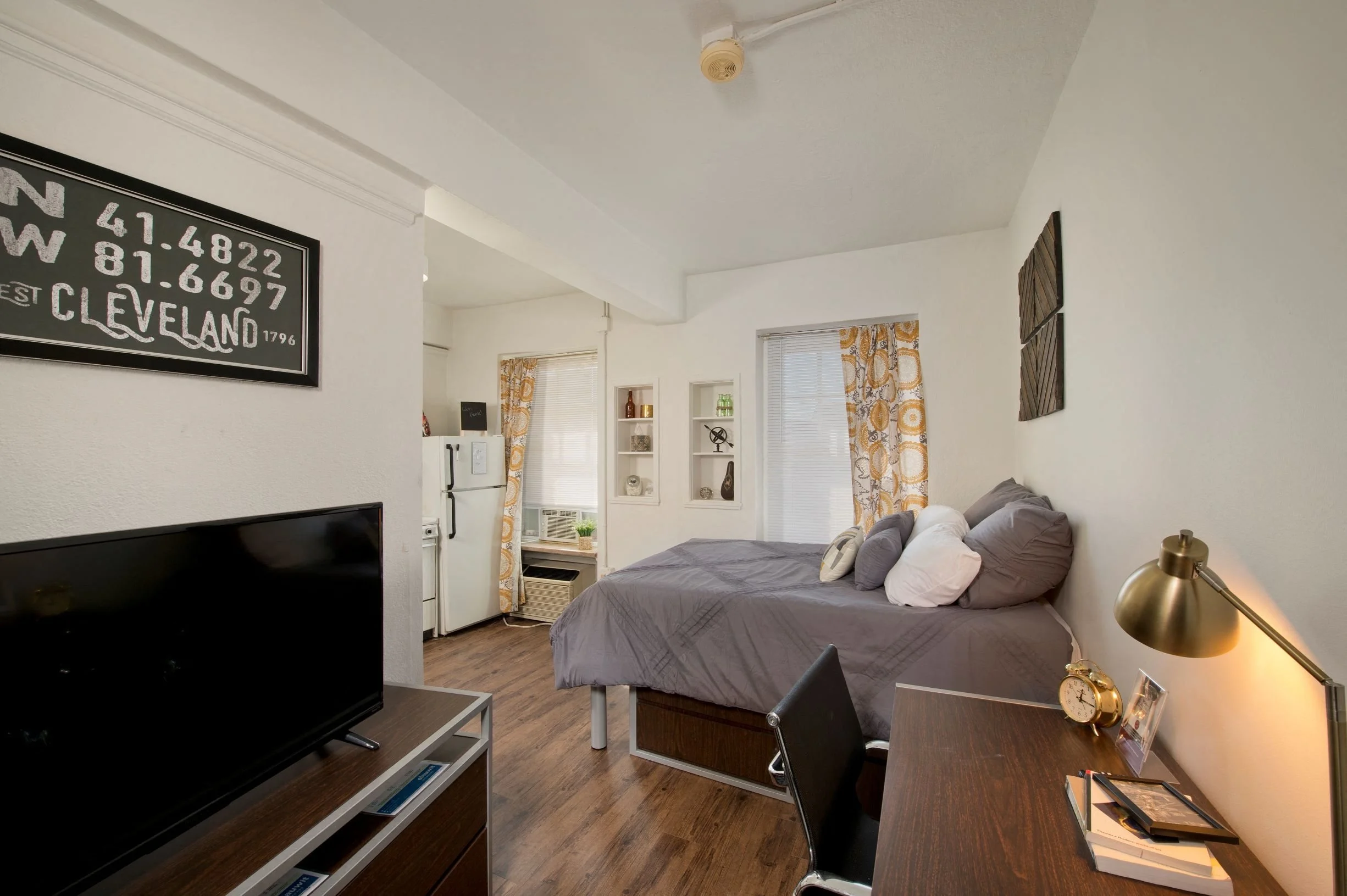 A cozy studio apartment featuring a bed, a desk, a television, and a small kitchenette with a refrigerator.