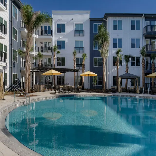 A modern apartment complex featuring a swimming pool with lounge chairs and umbrellas, surrounded by palm trees.