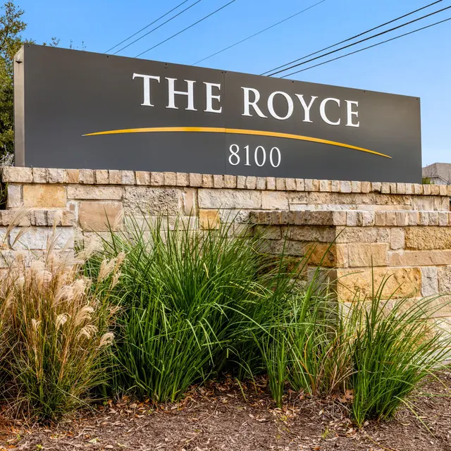 A sign for The Royce, featuring modern typography with the address 8100, set against a stone wall and surrounded by green plants.