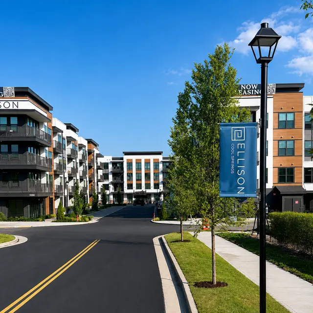 A modern apartment complex with two buildings, featuring balconies and a landscaped path leading to the entrance. A blue banner reading 'Ellison' is visible along with a street lamp and greenery.