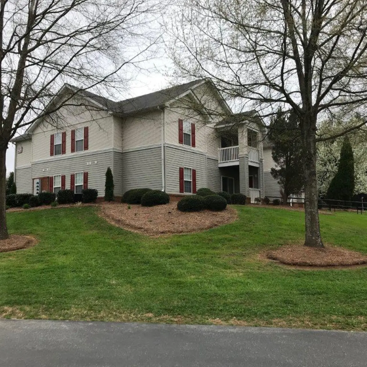 A two-story house with gray siding and red shutters, situated on a grassy hill surrounded by trees. The yard has landscaped mulch beds and a driveway leading up to it.