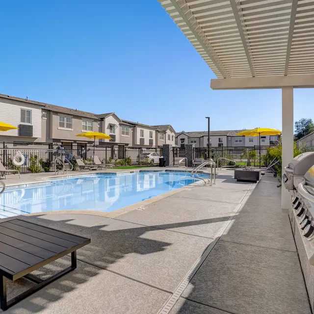 Sunny Pool Area in Apartment Complex A sunny pool area with lounge chairs, umbrellas, and a barbecue grill, surrounded by apartment buildings.