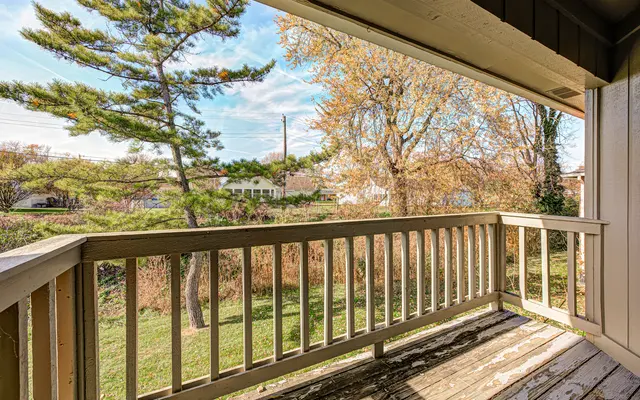A view from a balcony overlooking a green lawn with scattered trees and neighboring houses in the background under a partly cloudy sky.
