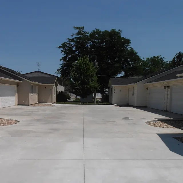 A view of a driveway surrounded by single-story houses, with a large tree in the middle and blue sky above.