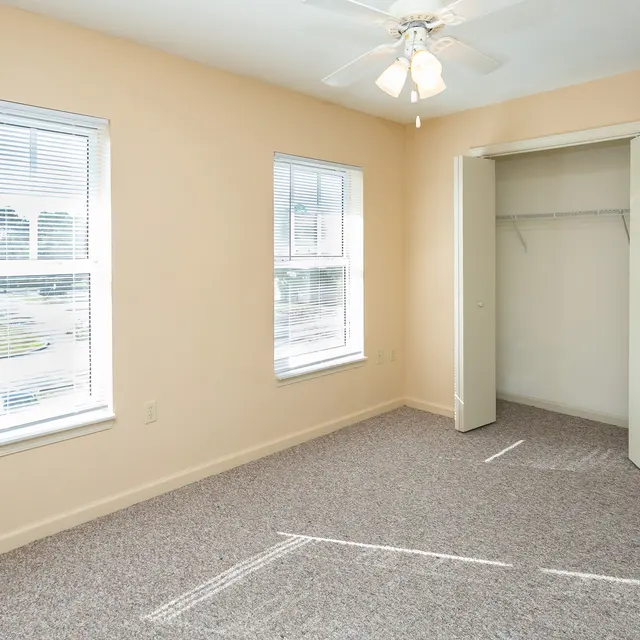 A bright, empty bedroom featuring two windows with blinds and a ceiling fan. The walls are painted a soft beige color, and the floor is covered in light carpet. A closet with an open door is visible in the corner.