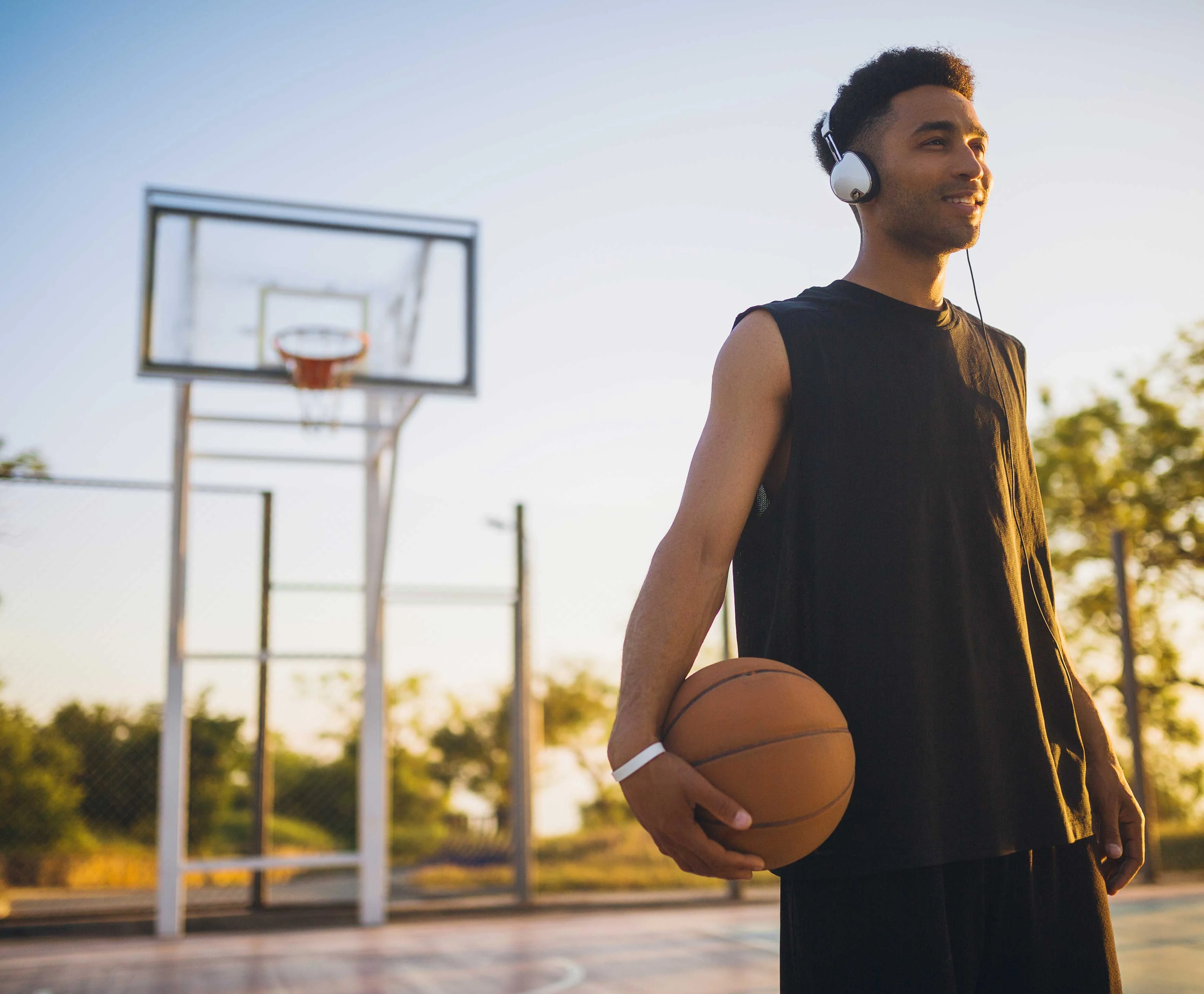 A young man wearing headphones stands on a basketball court at sunset, holding a basketball.