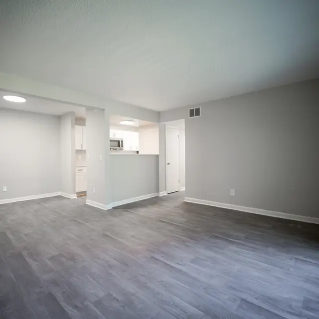 A spacious empty living room with light grey walls and wood-like flooring. The room has a doorway leading to a kitchen area visible in the background, with natural light coming from a ceiling fixture and large windows.