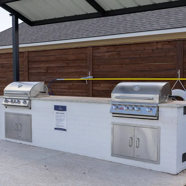 Outdoor grilling area with two stainless steel grills and a stone countertop, surrounded by landscaped greenery.
