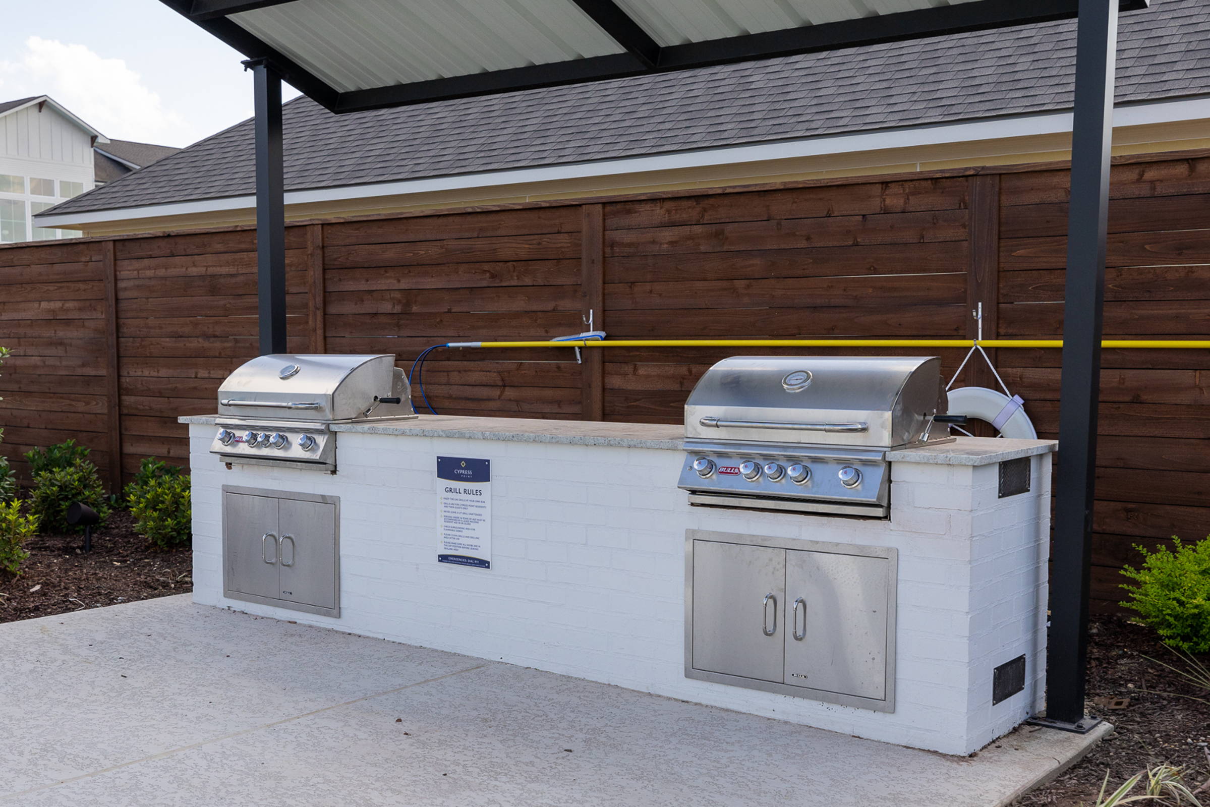 Outdoor grilling area with two stainless steel grills and a stone countertop, surrounded by landscaped greenery.