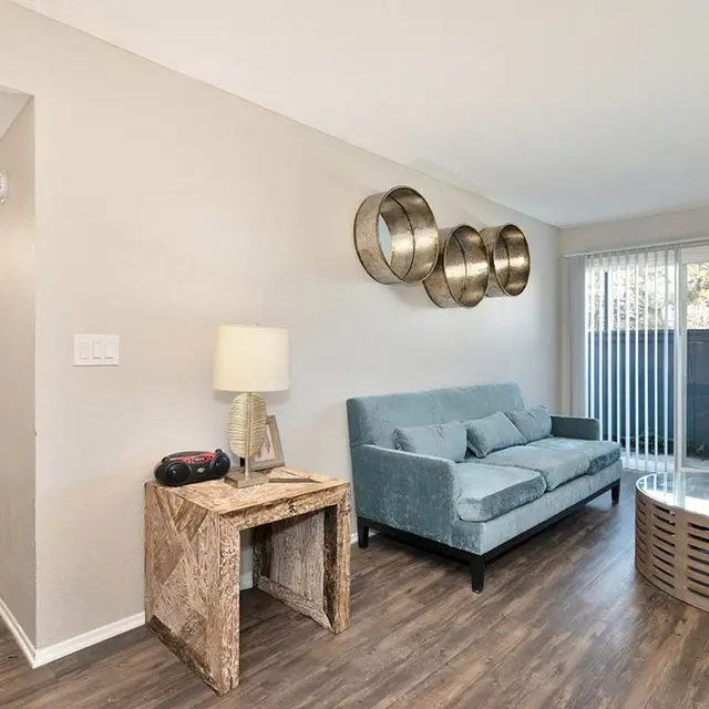 A light and airy living room featuring a blue couch, a round glass coffee table, and a lamp on a woven side table. A sliding glass door provides access to an outdoor area, and there are decorative wall hangings above the couch.