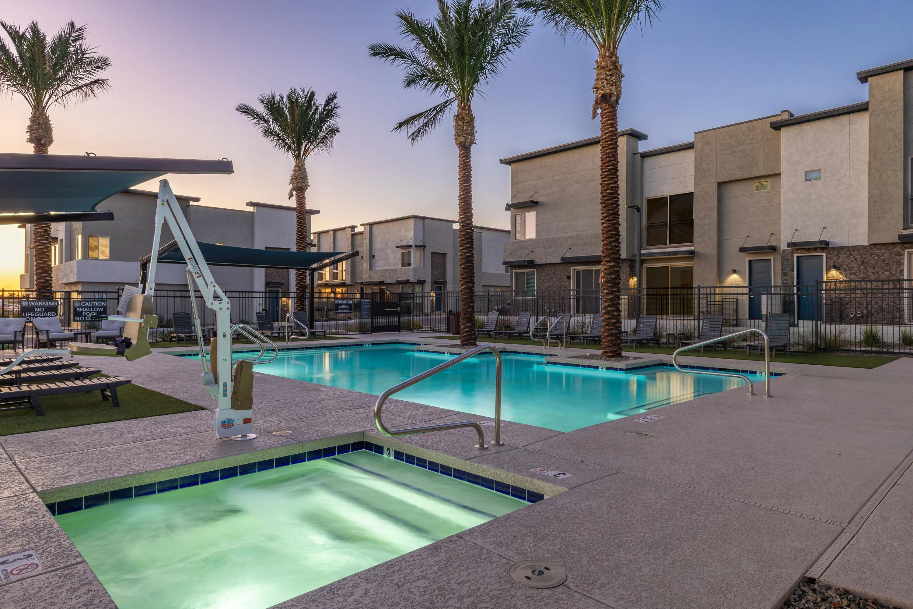A tranquil pool area featuring two swimming pools surrounded by palm trees and lounge chairs during sunset.