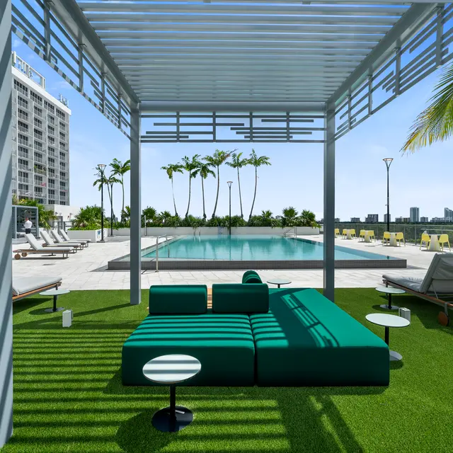 A contemporary lounge area with a green sectional sofa and small tables, overlooking a swimming pool surrounded by lounge chairs and palm trees, under a slatted sunshade.