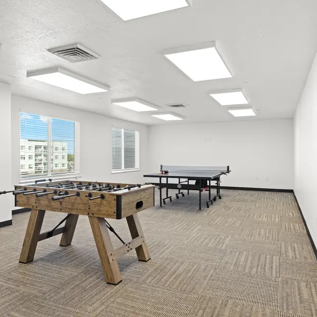 An empty game room featuring a foosball table and a ping pong table, with bright overhead lights and large windows letting in natural light.
