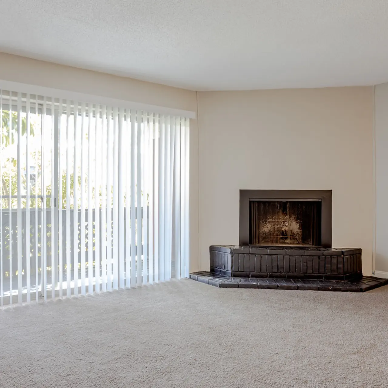A spacious living room featuring beige walls and carpet, a fireplace with a dark hearth, and large sliding glass doors with vertical blinds letting in natural light.