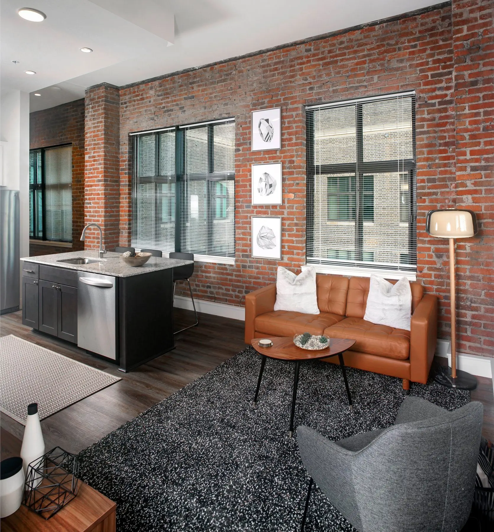 A modern loft interior featuring exposed red brick walls, large windows, and stylish furniture. The seating area includes a brown leather sofa with white cushions, a round coffee table, and a gray armchair on a dark rug. Next to the sofa, there is a modern floor lamp. The kitchen area showcases a dark island with stainless steel appliances.