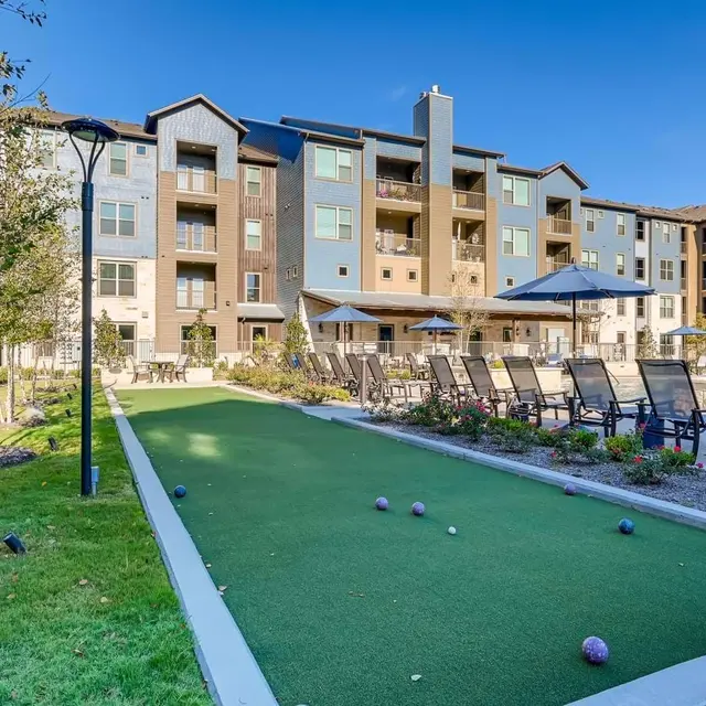 A well-maintained lawn area with a synthetic green bowling lane in front of a modern apartment building, featuring several lounge chairs and a patio area.