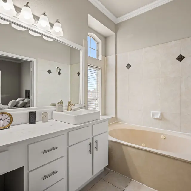 Modern Bathroom Design A modern bathroom featuring a large bathtub next to a white vanity with a sink and a mirror. The walls are painted light gray, and there are decorative tiles in the bathtub area. A window provides natural light, and a clock is placed on the vanity.