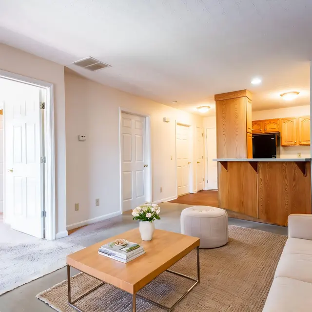 A cozy living room with a small wooden coffee table, a light-colored sofa adorned with cushions, and a decorative plant. Natural light fills the room, and an adjacent kitchen area can be seen in the background. Two open doors lead to other rooms, while a small stool and flowers on the table add decorative touches.