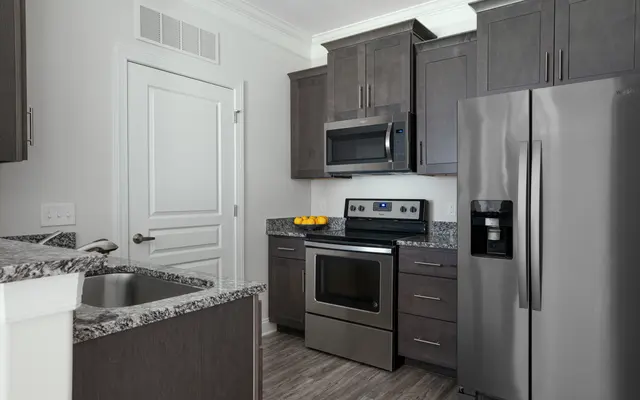 A brightly lit kitchen area with steel microwave, oven, and refrigerator. Surrounded by stained wood cabinets and granite countertops.