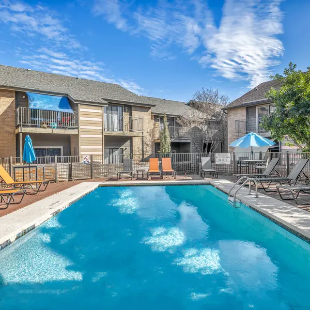 A sparkling swimming pool surrounded by lounge chairs, 2 umbrellas, trees, and 2-story apartment buildings.