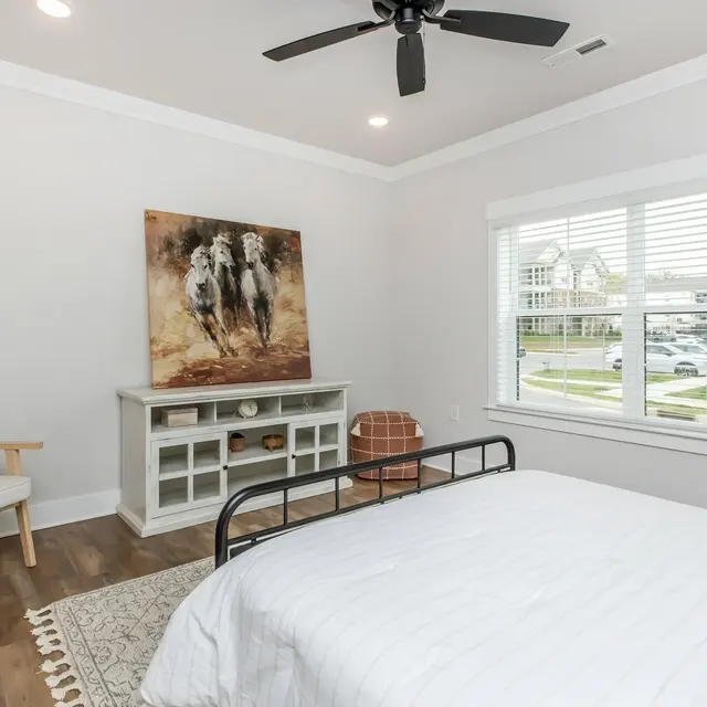 Cozy bedroom featuring a bed, wooden chair, artwork on the wall, and a window with blinds, creating a bright, inviting space.