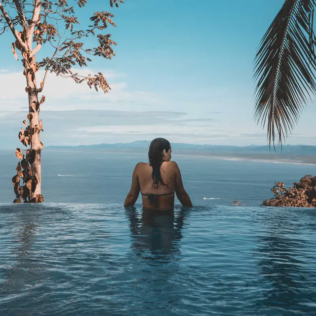 A person stands in an infinity pool overlooking a serene ocean landscape, with palm trees and clear blue skies.