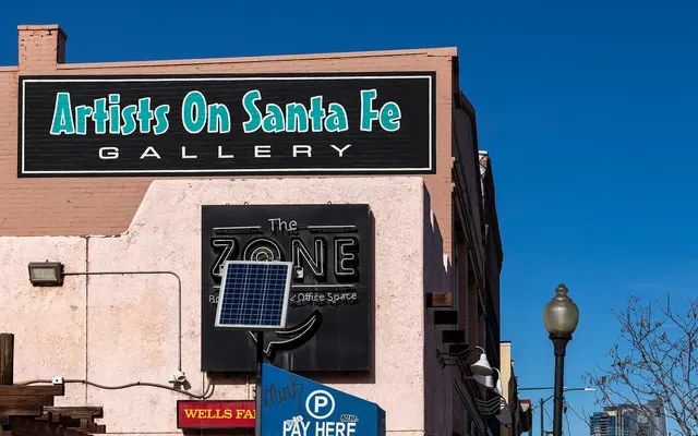 A building facade featuring a sign for 'Artists On Santa Fe Gallery' along with another sign for 'The Zone'. The scene is bright and clear, showcasing a sunny day with blue skies.