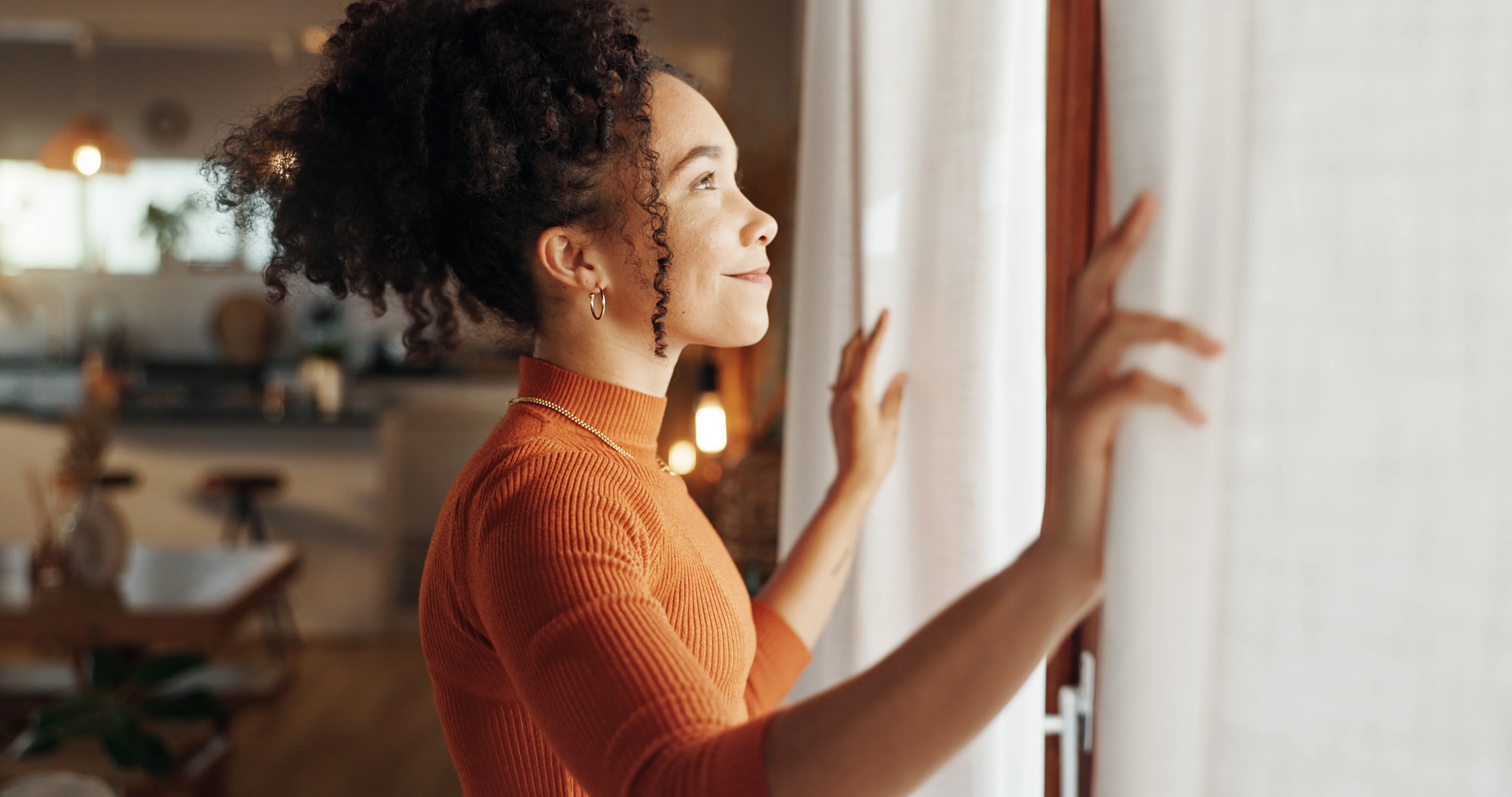 Contemplation by the Window A woman with curly hair stands by a window, holding back curtains and gazing outside with a serene expression. She wears an orange turtleneck sweater, and the background shows a softly lit room with a kitchen area visible.