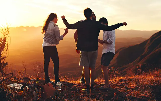 Group of friends enjoying a sunset on a mountain, dancing and laughing together in a scenic outdoor setting.