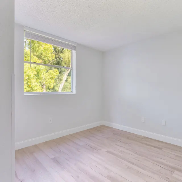 A minimalistic empty room with light-colored walls and wooden flooring. A window with natural light filtering in from the outside displays green trees.