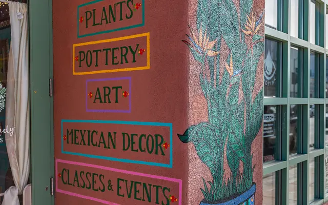 ARTISANALLY CRAFTED Colorful storefront signage with text highlighting offerings such as plants, pottery, art, Mexican decor, and classes & events, alongside a decorative mural featuring cacti.