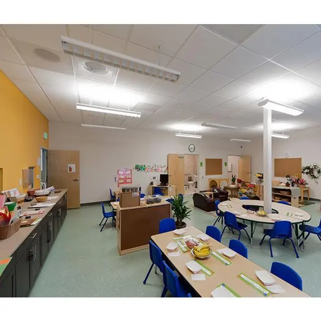 A bright and spacious classroom featuring multiple tables surrounded by blue chairs. One side has cabinets filled with learning materials, and another area shows small play stations. The walls are painted in warm colors, and there is ample overhead lighting.