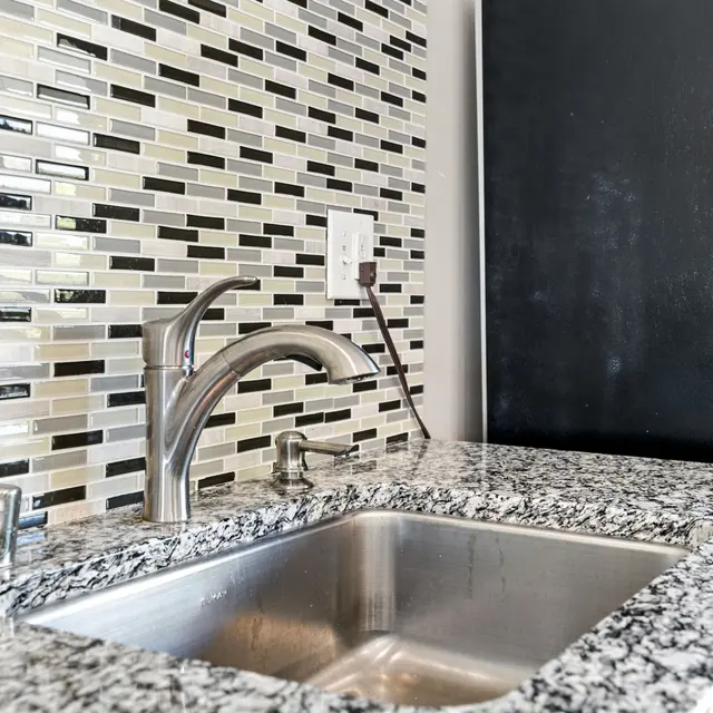 A modern kitchen sink with a stainless steel basin on a granite countertop, featuring a sleek faucet and a backsplash of patterned tiles in black, white, and green tones.