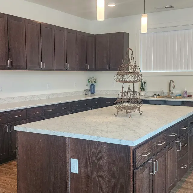 Canyon Terrace A modern kitchen featuring dark wooden cabinets, a large central island with a white countertop, and a black refrigerator. Natural light streams in through a window, illuminating the space.