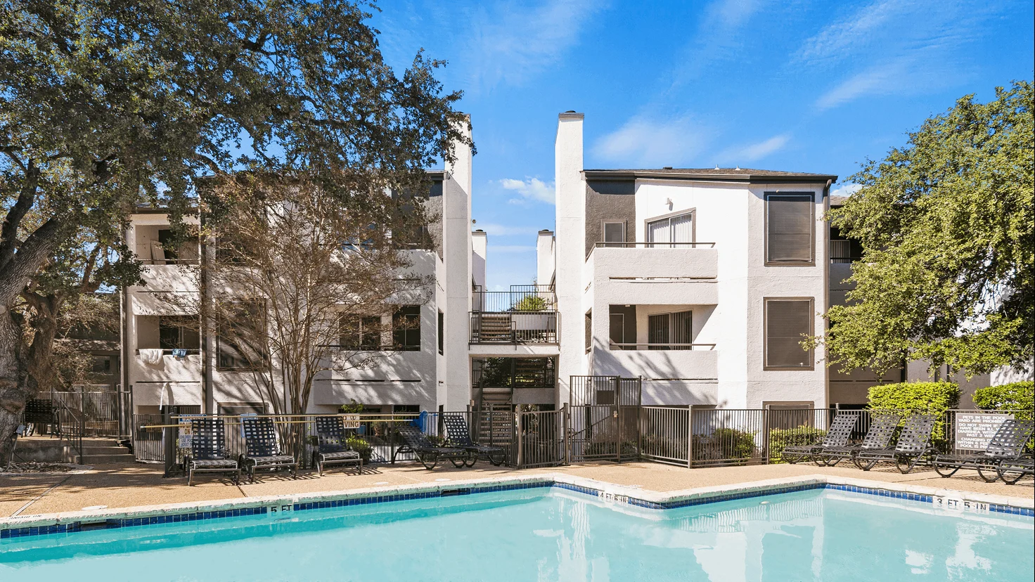 View of an apartment complex with a swimming pool in the foreground and tall trees nearby.