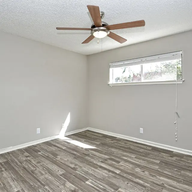 An empty room with gray walls, wood flooring, a ceiling fan, and a window with a pull-down blind.
