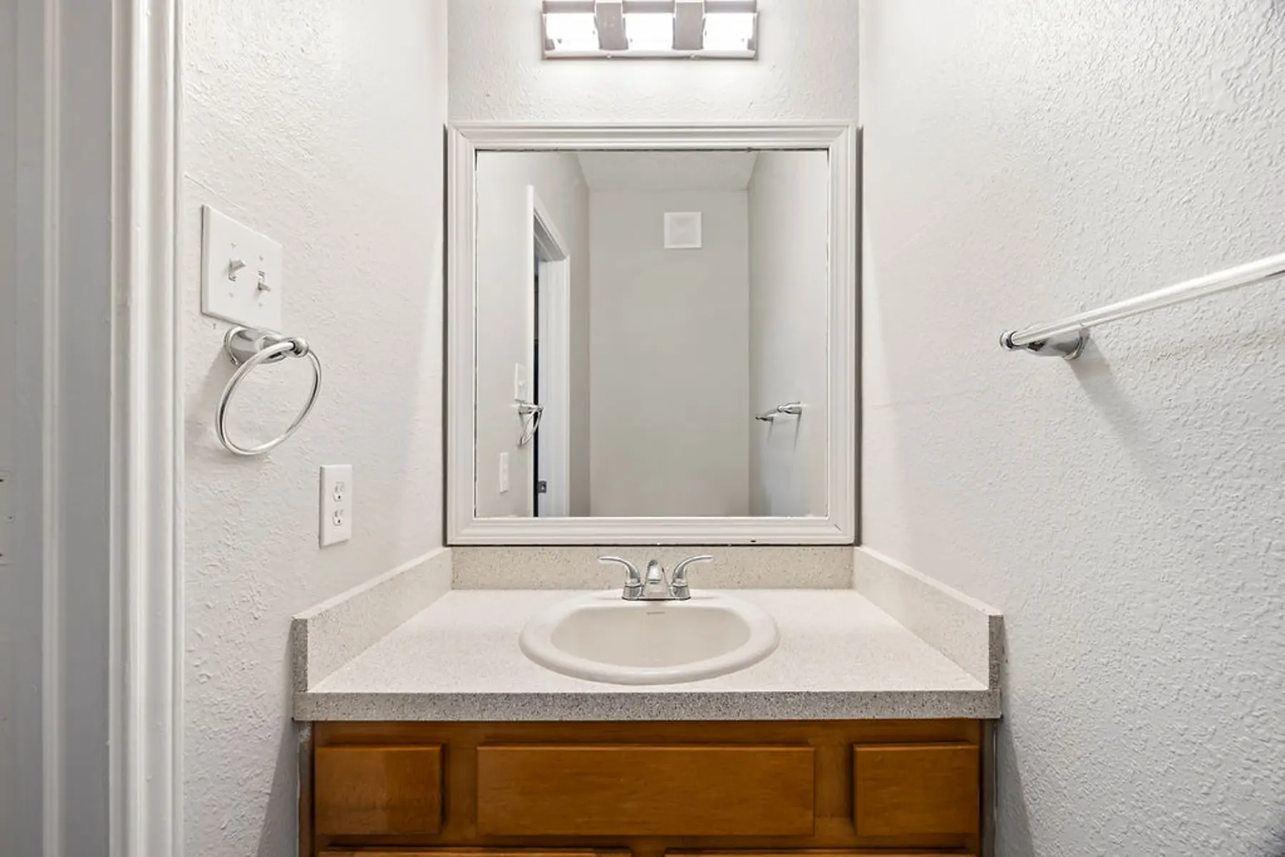 A clean and simple bathroom vanity with a sink, a mirror, and a light fixture above it. The walls are painted white, and the cabinet below is wooden.