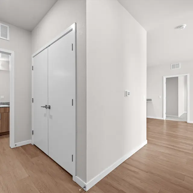 A spacious and modern hallway in an apartment, featuring light hardwood flooring, white walls, and an open layout. A door in the foreground leads to a closet, while a view of a bathroom with a countertop and mirror is visible on the left. In the background, another room is partially visible.