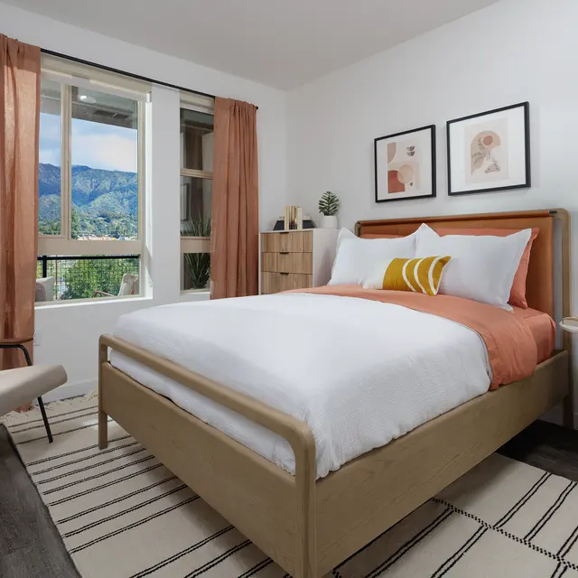 A cozy modern bedroom featuring a bed with white linens and an orange accent throw, complemented by a stylish chair, decorative wall art, and large windows revealing a mountainous view.