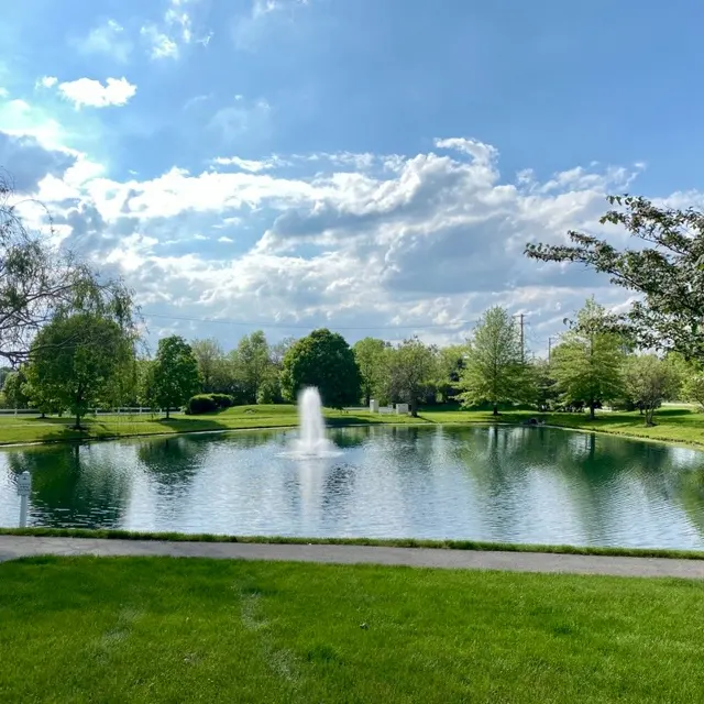 A serene park landscape featuring a pond with a fountain at its center, surrounded by green grass and trees under a partly cloudy sky.