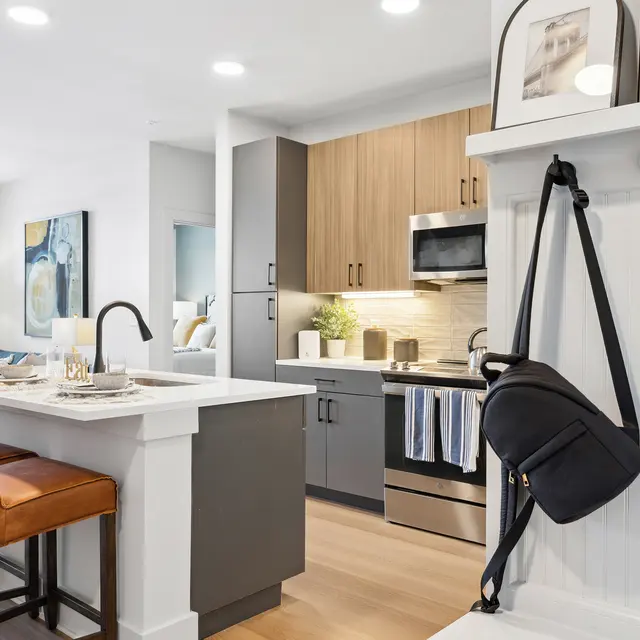 A modern kitchen with wooden and gray cabinetry, a stylish island with bar stools, and contemporary appliances. The area is bright with natural light and decorative elements are present.