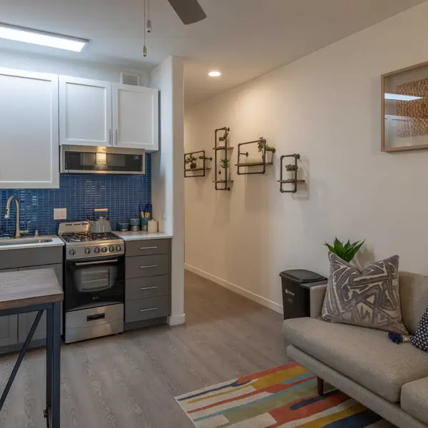 A modern kitchen and living room layout featuring a gray sofa, a colorful area rug, and a kitchen with white cabinets and a blue tiled backsplash.