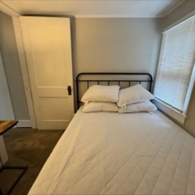 A cozy bedroom featuring a metal frame bed with white bedding and pillows, a bedside lamp on a wooden table, and a window with blinds letting in natural light. A closet door is also visible in the background.