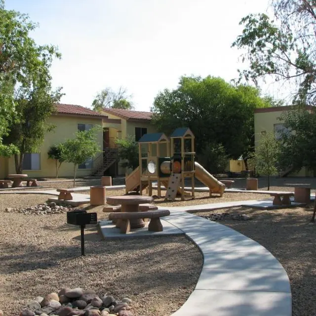Community Playground Area A playground area surrounded by trees and apartment buildings, featuring a wooden play structure, picnic tables, and a winding pathway made of concrete.