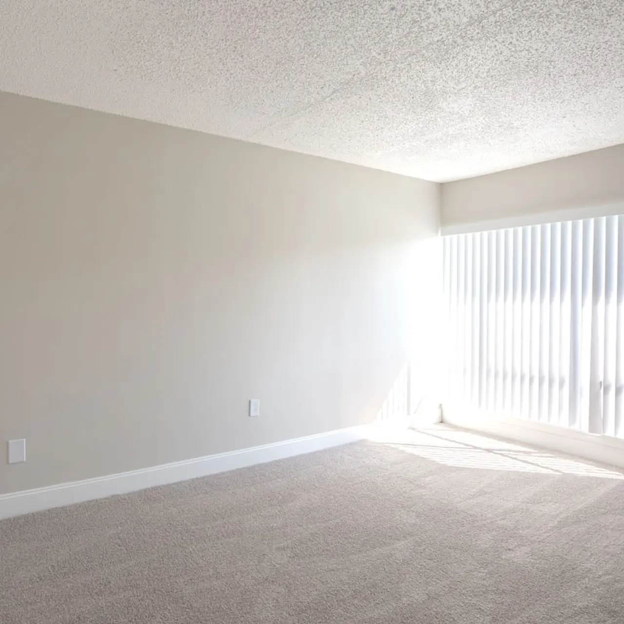 A spacious, empty room with light gray walls and a large window covered by vertical blinds, allowing natural light to fill the space.