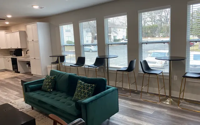 A modern living room featuring a green velvet sofa with decorative pillows, wood flooring, and tall bar stools beside large windows.