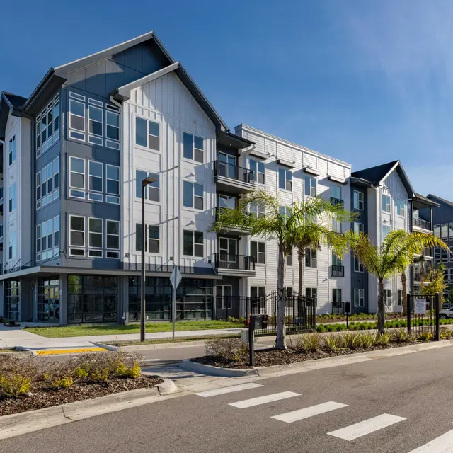 A modern apartment building with a mix of gray and white exteriors, surrounded by palm trees and landscaped areas. The street in front is lined with parked cars and has pedestrian crosswalks.